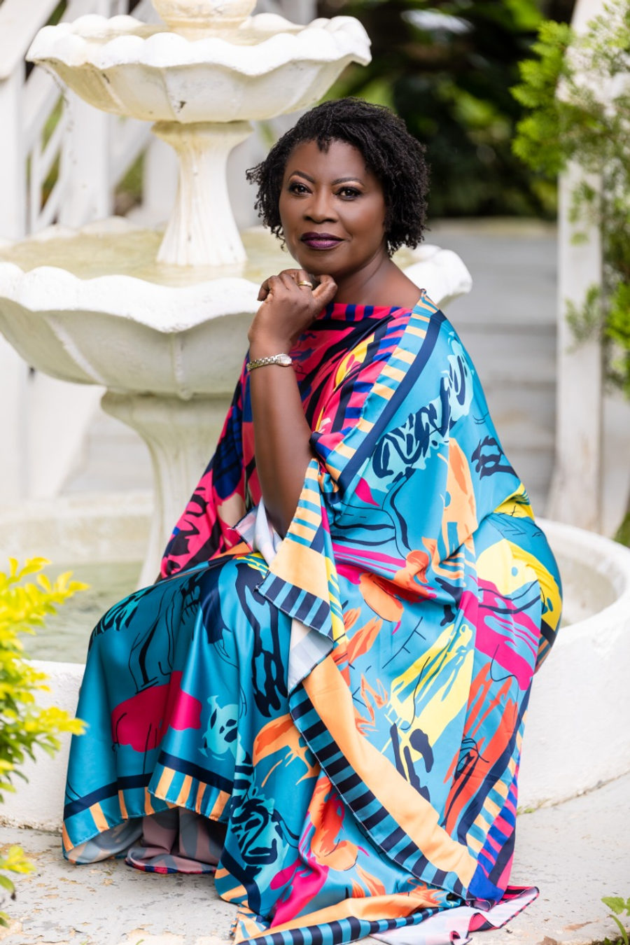 Ann-Marie Blaise seated beside a white garden fountain, wearing a bright patterned dress